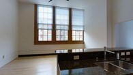 This single-floor space showcases a thoughtfully designed interior that blends functional and aesthetic elements. The kitchen area is anchored by sleek, dark granite countertops with a rich, speckled finish that adds depth and visual interest. The countertops extend along the workspace, providing ample surface area for meal preparation and daily use. A ceiling fan overhead aids in air circulation throughout the space.

Natural light is a defining feature, courtesy of three large, multi-pane windows framed in warm wood trim. The windows are fitted with roller shades that offer adjustable light control while maintaining clean sightlines. Views through the windows reveal a neighboring brick building, adding an urban character to the setting.

The flooring is light-toned hardwood, lending warmth and continuity throughout the open layout. The walls are finished in crisp white, creating a bright and airy backdrop that complements the darker countertops and wood accents. Electrical outlets are conveniently positioned along the counter backsplash. Overall, the space balances practical functionality with clean, modern design details.
