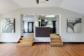 This single-floor space is thoughtfully designed for gatherings and events, offering a flexible, open layout across several interconnected areas. Warm hardwood-style flooring runs throughout, complemented by crisp white walls and bold black trim accents that lend a clean, modern aesthetic.

The main gathering hall is spacious and airy, featuring soaring vaulted ceilings with ceiling fans and recessed lighting that illuminate the room evenly. Large windows invite natural light and views of surrounding greenery, while a wall-mounted flat-screen television adds a functional focal point. An adjoining multi-level area connects seamlessly via wide wooden steps, creating distinct zones within the open floor plan.

The kitchen is fully equipped with dark cabinetry, granite-style countertops, pendant lighting, stainless steel appliances including a refrigerator and microwave, and a sink — all accessible via a raised platform with its own set of steps. A dark-paneled breakfast bar provides an informal service counter.

A separate dining or breakout room offers additional versatility, furnished with round white pedestal tables and stackable chairs. Black-framed French doors and windows brighten the space and provide outdoor access. A further open room with stacked folding chairs offers flexible extra capacity for events of varying sizes. Together, these areas make the space well-suited for meetings, receptions, or community gatherings.