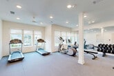 Located at 6101 Richmond Avenue in Houston, Texas, this fitness center offers a well-equipped exercise space designed for a comprehensive workout experience. The single-floor facility features a clean, modern aesthetic with crisp white walls, recessed lighting, and neutral carpet throughout, creating a bright and welcoming environment.

The main workout area is generously proportioned, accommodating a full range of exercise equipment including multiple treadmills, an elliptical machine, and a stationary bike for cardio training. A dedicated free weights section features a complete rack of kettlebells and dumbbells of varying sizes, along with adjustable workout benches for strength training. Floor-to-ceiling mirrors line one wall, enhancing the sense of space and allowing users to monitor their form during exercise.

Structural columns are integrated throughout the space, adding an architectural element while defining different workout zones. A separate anteroom adjacent to the main floor includes a built-in cubby shelving unit, providing convenient storage for personal belongings. Large windows with roller shades allow natural light to fill the cardio area, offering outdoor views of what appears to be a pool and surrounding landscaping. The space is accessed through glass-paneled doors, contributing to its open and airy feel.