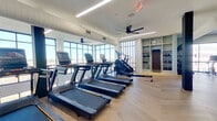 This thoughtfully designed fitness center offers a spacious, single-level layout with a generous floor area ideal for a full range of exercise activities. The space is divided into distinct functional zones, each crafted with both aesthetics and performance in mind.

The cardio zone is lined with a row of premium treadmills and elliptical machines positioned directly in front of large floor-to-ceiling windows, providing expansive outdoor views and abundant natural light that energize the workout experience. Overhead, sleek linear LED lighting and modern ceiling fans maintain a comfortable, well-ventilated atmosphere.

A dedicated cycling area houses smart stationary bikes equipped with interactive screens, set against a backdrop of additional large windows that continue the open, airy feel throughout the space. Nearby, a well-stocked equipment rack organizes stability balls, weights, and accessories, while built-in shelving along a sage-green accent wall offers additional storage for fitness gear.

An open studio area features a striking herringbone-patterned hardwood floor, providing an inviting surface for yoga, stretching, or group fitness classes. This zone is adorned with large abstract art pieces and framed by elegantly arched, backlit mirrors with integrated ballet barres — a refined architectural detail that adds both function and visual sophistication. TRX suspension trainers and a flat workout bench round out the strength and functional training options available throughout the facility.