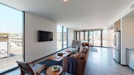 This single-floor urban residence showcases a distinctly modern architectural design, characterized by polished concrete floors, exposed concrete columns, and expansive floor-to-ceiling windows that frame sweeping cityscape views throughout. Natural light permeates the entire space, creating a bright, airy atmosphere.

The open-concept living area features a wall-mounted television and generous seating, flowing seamlessly into a dedicated dining area appointed with a full-size table and chairs. The dining space is anchored by a striking folding glass door system that opens to the outdoors, further connecting the interior to urban views beyond.

The kitchen is efficiently designed with warm wood cabinetry, a blue mosaic tile backsplash, stainless steel appliances including a range and microwave, and ample counter space. A hallway leads to a secure entry door.

The bedroom offers a tranquil retreat with continued concrete flooring, an exposed concrete accent wall, large windows overlooking the city, and a barn-style sliding door opening to a closet. The bathroom features bold geometric tile work, a glass-enclosed shower, open wood shelving, and a wall-mounted vanity, completing this refined and cohesive urban living space.