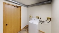 This single-floor laundry room is a practical, no-frills utility space designed for efficient everyday use. The room features a top-loading washer and dryer pair positioned along the wall, with dedicated hookups including water supply lines and electrical outlets conveniently accessible nearby. Exposed metallic ductwork runs prominently along the upper walls and ceiling, giving the space an industrial character while serving essential ventilation functions. The walls are finished in a neutral off-white tone, creating a clean and bright environment supported by overhead fluorescent lighting. Underfoot, durable stone-patterned tile flooring adds resilience suited to a high-use utility area. A smoke detector is mounted on the wall, and a posted notice is visible, indicating shared or managed use of the space. A wood-framed door with a door closer provides access to the room. The overall layout is straightforward and functional, with sufficient space to move around the appliances comfortably. The room is well-suited to handle laundry needs efficiently, with all essential utilities clearly in place.