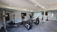 This fitness center is thoughtfully organized into distinct workout zones, offering a comprehensive exercise environment across a single floor. The main cardio area features multiple elliptical trainers and treadmills arranged on durable, cushioned tile flooring, with a wall-mounted flat-screen TV and recessed lighting overhead. Structural beams and a coffered ceiling detail add architectural character, while large glass double doors flood the space with natural light and provide views of a lush outdoor courtyard. A dedicated cycling studio is separated by full-length glass partition walls, housing a row of spin bikes facing a large wall-mounted television. Full-wall mirrors amplify the sense of space and allow users to monitor their form. Skylight panels in the cycling room provide additional natural illumination. A strength and free weights alcove features a multi-station cable machine, a tiered dumbbell rack, and an adjustable weight bench, also appointed with mirrored walls and skylight panels. The neutral gray and white color palette throughout creates a clean, modern aesthetic. The facility is well-lit with a combination of recessed downlights and natural light sources, maintaining an open and motivating atmosphere suited for a variety of fitness routines.