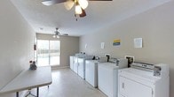This shared laundry facility is a practical and functional space designed to serve the needs of its users efficiently. The room is outfitted with multiple full-sized washing machines and dryers arranged in a neat row along the walls, providing ample capacity for simultaneous use. Payment kiosks are conveniently integrated into the setup, streamlining the laundry process. The space features a textured ceiling with two ceiling fans that promote air circulation and comfort throughout the room. A large window fitted with horizontal blinds allows natural light to fill the space while offering a view of an outdoor area, likely a pool or courtyard. The flooring is a smooth, easy-to-clean surface well-suited for a high-traffic utility space. Neutral-toned walls contribute to a clean and uncluttered atmosphere. Informational notices are posted on the walls, offering guidance on the use of the machines and facility rules. The layout is open and spacious, allowing multiple users to move around comfortably. Overall, the facility is a well-maintained, no-frills laundry space built for convenience and practicality.