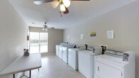 This shared laundry facility is a practical and functional space designed to serve the needs of its users efficiently. The room is outfitted with multiple full-sized washing machines and dryers arranged in a neat row along the walls, providing ample capacity for simultaneous use. Payment kiosks are conveniently integrated into the setup, streamlining the laundry process. The space features a textured ceiling with two ceiling fans that promote air circulation and comfort throughout the room. A large window fitted with horizontal blinds allows natural light to fill the space while offering a view of an outdoor area, likely a pool or courtyard. The flooring is a smooth, easy-to-clean surface well-suited for a high-traffic utility space. Neutral-toned walls contribute to a clean and uncluttered atmosphere. Informational notices are posted on the walls, offering guidance on the use of the machines and facility rules. The layout is open and spacious, allowing multiple users to move around comfortably. Overall, the facility is a well-maintained, no-frills laundry space built for convenience and practicality.
