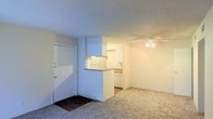 This single-floor unit presents an open and functional layout designed for comfortable everyday living. The living area is finished with wall-to-wall carpet, lending a warm and soft underfoot feel to the space. A ceiling fan is centrally positioned overhead, providing both light and air circulation throughout the room.

The kitchen is defined by a raised pass-through counter that separates it from the living area while maintaining an open connection between the two spaces. White cabinetry lines the kitchen walls, offering ample storage, and white appliances complement the clean, cohesive aesthetic. The countertop extends along the perimeter, providing functional workspace for meal preparation.

Near the entrance, the flooring transitions from carpet to a darker hard surface, creating a natural entryway zone just inside the front door, which features a standard white panel design with a brass-finished handle and deadbolt. The walls throughout are finished in a neutral tone, complemented by a textured ceiling that adds subtle visual depth. Overall, the space maintains a straightforward, practical character suited for a range of occupants.