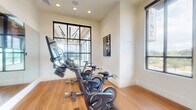 Located at 9901 W 58th Ave in Arvada, Colorado, this dedicated cycling studio is a well-appointed fitness space designed specifically for indoor cycling workouts. The room is outfitted with multiple Echelon stationary bikes, each equipped with individual monitors for on-demand or live class streaming, making it a fully functional spin studio.

The space features warm hardwood flooring throughout, which adds both durability and an inviting aesthetic. A large wall-mounted mirror spans a significant portion of one wall, visually expanding the room and allowing cyclists to monitor their form during workouts. Recessed ceiling lighting provides consistent, even illumination across the studio.

Natural light is a defining feature of this space, with multiple large, black-framed windows lining the walls and offering views of the surrounding outdoor greenery. The windows not only enhance the brightness of the room but also create an open, airy atmosphere that supports an energizing workout environment.

An arched doorway visible through the mirrored wall hints at a connection to an adjacent, larger fitness area. Cycling-themed wall art and informational signage add purposeful, motivational décor. The clean, neutral wall tones paired with the warm flooring create a modern yet welcoming studio environment suited for group or individual cycling sessions.