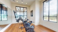 Located at 9901 W 58th Ave in Arvada, Colorado, this dedicated cycling studio is a well-appointed fitness space designed specifically for indoor cycling workouts. The room is outfitted with multiple Echelon stationary bikes, each equipped with individual monitors for on-demand or live class streaming, making it a fully functional spin studio.

The space features warm hardwood flooring throughout, which adds both durability and an inviting aesthetic. A large wall-mounted mirror spans a significant portion of one wall, visually expanding the room and allowing cyclists to monitor their form during workouts. Recessed ceiling lighting provides consistent, even illumination across the studio.

Natural light is a defining feature of this space, with multiple large, black-framed windows lining the walls and offering views of the surrounding outdoor greenery. The windows not only enhance the brightness of the room but also create an open, airy atmosphere that supports an energizing workout environment.

An arched doorway visible through the mirrored wall hints at a connection to an adjacent, larger fitness area. Cycling-themed wall art and informational signage add purposeful, motivational décor. The clean, neutral wall tones paired with the warm flooring create a modern yet welcoming studio environment suited for group or individual cycling sessions.
