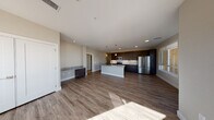 This single-floor residence showcases a thoughtfully designed open-concept layout where the kitchen serves as the centerpiece. The kitchen is outfitted with rich, dark espresso cabinetry extending to the ceiling, granite countertops, a full stainless steel appliance suite including a refrigerator, range, microwave, and dishwasher, and a white-paneled island with a granite surface and undermount sink. A white subway tile backsplash adds a clean contrast to the dark cabinetry, while pendant lighting above the island enhances the functional aesthetic. Wood-look plank flooring flows throughout the main living areas, providing a warm, cohesive feel. The open living area offers generous space with natural light filtering in through windows offering exterior views. The bedroom features plush carpet, a ceiling fan, a closet, and a built-in desk with granite countertop. The bathroom includes dark vanity cabinetry with granite countertops, an undermount sink, a large mirror, and a tub-shower combination with white tile surround. A dedicated laundry area houses a full-size washer and dryer with overhead dark cabinetry for additional storage. Neutral wall tones throughout create a clean, contemporary ambiance.