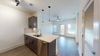 This single-floor space showcases a contemporary design with a cohesive blend of dark-toned cabinetry and light finishes throughout. The open-concept kitchen serves as a focal point, featuring a peninsula with a white quartz countertop, an undermount stainless steel sink, and pendant lighting suspended above. Dark espresso cabinetry with sleek hardware pairs with a subway tile backsplash, while stainless steel appliances—including a refrigerator, range, microwave, and dishwasher—complete the functional layout. A wine rack adds a distinctive storage detail above the upper cabinets.

The kitchen flows seamlessly into the living area, where wood-style plank flooring extends throughout and a ceiling fan provides overhead comfort. Natural light enters through multiple windows, including what appears to be a door with glass panels leading to an outdoor area.

The bathroom is thoughtfully designed with a full soaking tub and shower combination enclosed by white subway tile, a dark-framed vanity mirror, light countertops, a multi-bulb vanity fixture, and wood-style flooring that ties the space together cohesively.

The bedroom offers plush carpeting, a ceiling fan, and a window that brings in natural light, creating a calm and comfortable retreat.