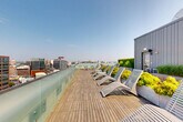 Located on Boylston Street in Boston, MA, this rooftop terrace offers an expansive outdoor retreat with sweeping panoramic views of the city skyline and waterfront. The deck is finished with warm wood planking throughout, creating a cohesive and inviting atmosphere across its generous open-air footprint.

The terrace is thoughtfully divided into distinct seating zones. One area features rows of slatted chaise lounges paired with large concrete planters overflowing with vibrant yellow-green shrubs and ornamental grasses, ideal for sunbathing and relaxation. A second zone offers a social gathering space with modern molded lounge chairs arranged in an open-facing configuration, well-suited for conversation and events. A third area provides additional lounge seating surrounded by lush greenery and tall ornamental grasses.

A sleek, modern rooftop structure clad in gray vertical metal panels anchors the space, providing interior access and housing mechanical systems. Glass panel railings line the perimeter, preserving unobstructed sightlines to Boston's urban landscape, including recognizable downtown towers, neighboring rooftops, and glimpses of the harbor. The overall design balances contemporary urban aesthetics with abundant greenery, creating a verdant rooftop oasis above the city.