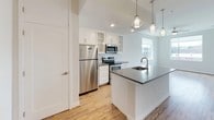 This 1-bedroom, 1-bathroom unit is designed with a clean, modern aesthetic and a thoughtfully open layout. The living area features light wood-style flooring, a ceiling fan with integrated lighting, and large windows that invite natural light and offer views of the surrounding urban streetscape. The bright, neutral palette throughout creates an airy, welcoming ambiance.

The kitchen is a standout feature, centered around a peninsula island with a dark gray countertop, an undermount sink, and elegant pendant lighting above. White shaker-style cabinetry — including glass-front upper cabinets — pairs with a subway tile backsplash and a full suite of stainless steel appliances, including a refrigerator, electric range, and built-in microwave. Wood-style flooring continues seamlessly through this space.

The bedroom offers plush carpeting, a ceiling fan, and a well-sized window with natural light and exterior views, providing a comfortable and restful retreat.

The bathroom is finished in crisp white tones and includes a full tub-shower combination with a curved shower rod and recessed overhead lighting, maintaining the unit's consistent, clean design language throughout.