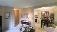 This single-floor space offers a warm and inviting layout with a seamless flow between its main living areas. The living room features neutral-toned carpeting and beige walls, creating a cozy atmosphere complemented by traditional décor, including framed botanical artwork and a dark wood console table. Candlestick accents and a coffee table add character to the space.

An open passageway connects the living room to the kitchen and dining area, allowing for easy visibility throughout. The kitchen is equipped with white cabinetry, a gas range, and a range hood, reflecting a functional and clean design. Adjacent to the kitchen, the dining area features a dark wood high-top table paired with matching bar-height chairs, illuminated by an elegant pendant light fixture that anchors the space.

A hallway visible from the living room leads further into the unit, lined with what appears to be a louvered closet door, suggesting additional storage. The textured ceiling and warm ambient lighting throughout contribute to the overall comfortable and homey feel of this well-connected, single-level space.