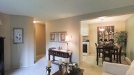 This single-floor space offers a warm and inviting layout with a seamless flow between its main living areas. The living room features neutral-toned carpeting and beige walls, creating a cozy atmosphere complemented by traditional décor, including framed botanical artwork and a dark wood console table. Candlestick accents and a coffee table add character to the space.

An open passageway connects the living room to the kitchen and dining area, allowing for easy visibility throughout. The kitchen is equipped with white cabinetry, a gas range, and a range hood, reflecting a functional and clean design. Adjacent to the kitchen, the dining area features a dark wood high-top table paired with matching bar-height chairs, illuminated by an elegant pendant light fixture that anchors the space.

A hallway visible from the living room leads further into the unit, lined with what appears to be a louvered closet door, suggesting additional storage. The textured ceiling and warm ambient lighting throughout contribute to the overall comfortable and homey feel of this well-connected, single-level space.