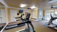 This single-floor fitness space offers a clean, functional layout designed to support an active lifestyle. The room is finished with warm yellow walls and white crown molding, lending a polished, welcoming atmosphere that distinguishes it from a typical gym setting. Carpeted flooring provides cushioning underfoot throughout the workout area, while a large multi-pane window along one wall invites abundant natural light and offers views of surrounding greenery, creating an open, airy feel.

The space is equipped with multiple cardio machines, including treadmills and an elliptical trainer, thoughtfully arranged to allow comfortable use. A wall-mounted flat-screen television is positioned for easy viewing during workouts, accompanied by additional wall-mounted accessories nearby. A ceiling fan with a central light fixture ensures adequate air circulation and overhead illumination.

A built-in white shelving and cubby unit with a lower bench runs along one wall, providing practical storage and a convenient spot for belongings. Coat hooks mounted beside the shelving add further organizational utility. The overall design balances fitness functionality with residential comfort and attention to detail.