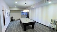 This single-floor recreational space is thoughtfully designed to offer a polished and inviting environment for leisure activities. The centerpiece of the room is a full-size billiards table finished in a sleek black frame with a light grey felt surface, perfectly positioned beneath a set of contemporary rectangular pendant lights that cast a warm, focused glow over the playing area. The room features neutral-toned walls accented with subtle horizontal striping, lending a refined, modern aesthetic throughout. Patterned carpet tiles in a grey hue cover the floor, adding texture and a sense of sophistication underfoot. Recessed ceiling lighting complements the pendant fixtures, ensuring the space is well-lit from every angle. An adjacent area visible in the background hints at additional amenities or a connected lounge space, suggesting a broader entertainment zone. The open layout provides ample room to move freely around the table, making it well-suited for both casual play and social gatherings. Overall, the space strikes a balance between functionality and modern design, offering a comfortable and stylish setting for recreation.