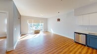 This single-floor residence at CiTi Village in Linden, New Jersey offers a clean, contemporary design throughout. Warm wood-style flooring flows consistently across the main living areas, complementing the neutral gray-toned walls and bright white trim that define the interior aesthetic.

The open-concept living area is generously proportioned, bathed in natural light through large windows that offer outdoor views. A stylish pendant light fixture adds a decorative focal point to the space. The living area connects seamlessly to the kitchen and entry foyer, creating an efficient and welcoming flow.

The kitchen is a standout feature, showcasing two-tone cabinetry with vibrant teal lower cabinets and crisp white upper cabinets. A full suite of stainless steel appliances—including a gas range, over-the-range microwave, refrigerator, and dishwasher—provides modern functionality alongside a deep single-basin sink with ample counter space.

The bedroom is a comfortable retreat with dual closets offering generous storage. A window allows natural light to fill the room. The unit includes one-and-a-half bathrooms, both finished with large-format neutral floor tiles, floating dark wood vanities with white countertops, framed mirrors, and bright vanity lighting, delivering a polished, modern aesthetic.