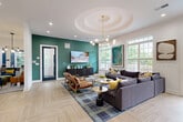Located at 138 Greison Trail in Newnan, Georgia, this single-floor community clubhouse offers a well-appointed and thoughtfully designed gathering space. The interior is anchored by a generous open lounge area showcasing a signature coffered circular ceiling detail, an eye-catching multi-globe brass chandelier, and a bold teal accent wall. Light wood-look tile flooring laid in a herringbone pattern flows throughout the space, lending warmth and visual continuity. Large windows and black-framed French doors flood the interior with natural light while framing views of lush greenery outside. A built-in wet bar with navy cabinetry and decorative tile backsplash adds a refined entertaining element to the lounge. The conference room features a substantial live-edge wood table, teal upholstered seating, a wall-mounted display screen, and a dramatic full-wall artistic mural, all illuminated by sleek brass pendant lighting. The leasing office area is equally polished, with rich walnut desking, contemporary pendant fixtures, and curated abstract artwork throughout. The welcoming entry foyer features an arched transom above the French doors, a console table vignette, and cognac leather accent chairs, setting an inviting tone from arrival.