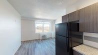Located in Cincinnati, OH (45219), this single-floor unit offers a functional and modern living layout. The open main living area is characterized by light wood-style luxury vinyl flooring, white walls, and a textured ceiling, creating a clean and bright atmosphere. A large window floods the living space with natural light while framing views of the surrounding cityscape and green landscape, enhancing the sense of openness throughout.

The kitchen is well-appointed and runs along a single wall, featuring dark wood-toned cabinetry that extends to the ceiling for ample storage, granite-style countertops, and a full suite of black appliances including a refrigerator, electric range, built-in microwave, and dishwasher. A stainless steel sink and tiled backsplash complete the functional workspace.

The bathroom includes a granite-top vanity with an undermount sink, white square-tile walls, and a separate tub/shower combination, along with a walk-in closet with wire shelving conveniently located nearby.

Recessed lighting in the kitchen and entryway provides targeted illumination, while a wall-mounted HVAC unit in the living area ensures year-round climate control. The entry area is well-defined, offering a clear transition into the living space. Overall, the unit presents a practical, modern design suited for everyday living in an urban Cincinnati setting.