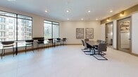 Located at 1320 Stanley St NE in Sugar Hill, GA, this contemporary single-floor space offers a thoughtfully designed lounge and meeting environment. The main area is anchored by a large conference-style table with upholstered chairs, complemented by a bar-height counter and seating along expansive windows that flood the room with natural light and offer views of the surrounding community. Light-toned terrazzo-style flooring flows throughout, lending the space a clean, polished aesthetic. The walls are adorned with curated abstract artwork, while warm gold-toned wall sconces provide accent lighting alongside recessed ceiling fixtures. A paneled feature wall serves as a striking focal point, framing a mounted display and adding architectural depth. A hallway connects to additional rooms, including a private office or meeting room accessible through a sliding barn door, as well as a glass-door enclosure and a utility area housing office equipment with cabinetry. The neutral color palette of warm whites, tans, and soft gray-green accents creates a professional yet welcoming ambiance, making this space well-suited for collaboration, community gatherings, or business functions.