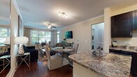 This single-floor residence offers a thoughtfully designed open-concept layout that seamlessly connects the living, dining, and kitchen areas, creating a cohesive and functional living environment. Rich hardwood-style flooring flows throughout the main living spaces, complemented by crown molding and neutral warm-toned walls that enhance the overall sense of elegance.

The living room is a comfortable and stylish retreat, featuring a ceiling fan with light fixture, large windows that invite abundant natural light and views of surrounding greenery, and ample space for both relaxing and entertaining. A wall-mounted flat-screen TV and an accent wall with large-format artwork add character to the space.

The kitchen is well-equipped with dark espresso cabinetry, granite-style countertops, stainless steel appliances including a refrigerator, range, and microwave, and a double-basin sink. A breakfast bar peninsula connects the kitchen to the dining area, where a round pedestal table provides an inviting space for meals.

The bathroom features dark cabinetry with a granite-topped vanity, a framed mirror, and a shower-tub combination with tiled surround. Additional highlights throughout the residence include louvered closet doors, a dedicated entry area with console storage, and an overall design that balances practicality with refined style.