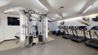This fitness center is a professionally designed workout facility that combines functionality with a clean, modern aesthetic. The space features smooth, light-toned ceilings fitted with recessed and linear LED lighting that bathes the entire floor in bright, even illumination, creating an energizing atmosphere for exercise.

At the forefront of the space stands a multi-functional cable and pulley training rig from Life Fitness, offering a wide range of resistance-based exercises including TRX suspension training. Adjacent to this, a dedicated stretching and floor exercise area is defined by a green rubber mat, with stability and medicine balls stored nearby for dynamic workout options.

Along the far wall, a row of treadmills is neatly arranged, each equipped with individual display screens for interactive workout tracking. The flooring throughout is a durable, speckled rubber gym surface designed to absorb impact and provide traction during training.

A standout design feature is the bold motivational mural on the accent wall, incorporating graphic lettering and an emblem that lends the space a strong team-oriented identity. A glass partition separates an additional exercise zone, adding depth and visual openness to the layout. The overall design prioritizes both performance and atmosphere, making this a comprehensive and inviting fitness facility.
