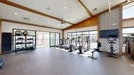 Located at 2500 East Marco Polo Road in Phoenix, AZ, this generously sized fitness center offers a well-appointed exercise space designed to support a full range of workout routines. The facility is characterized by soaring ceilings with exposed wood beam detailing and warm wood accents throughout, lending an upscale, modern aesthetic. Floor-to-ceiling windows and large sliding glass doors flood the interior with abundant natural light while offering views of the surrounding outdoor environment. The rubber-floored exercise area is organized to accommodate both cardio and strength training, housing a comprehensive lineup of treadmills, ellipticals, rowing machines, cable machines, free weights, benches, and multi-functional strength equipment. A decorative patterned tile accent wall serves as a focal point, complemented by wall-mounted TVs for entertainment during workouts. Hanging globe pendants and lush trailing plants add an inviting, biophilic touch to the high-volume space. Large ceiling fans provide climate comfort, while a staircase connects to an upper observation level. Sliding glass doors open to a private outdoor terrace featuring artificial turf, perfect for outdoor training or stretching. The overall ambiance is bright, airy, and motivating — a premium fitness environment suited for a wide variety of exercise preferences.