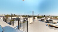 This expansive rooftop terrace offers a well-appointed outdoor experience with sweeping panoramic views of the surrounding cityscape, tree canopy, and distant mountain ranges under open skies. The deck is thoughtfully divided into multiple functional zones, each designed to accommodate different activities and group sizes. Overhead string lights are strung between dark metal posts, creating an inviting ambiance that transitions seamlessly from day to evening use. Several distinct seating clusters feature contemporary upholstered lounge sofas and chairs arranged around low tables, ideal for relaxed conversation or social gatherings. A dedicated dining area with a table and chairs provides a space for al fresco meals. Galvanized metal trough planters are distributed throughout, adding greenery and a modern industrial aesthetic. A sunbathing zone is equipped with chaise lounge chairs and side tables, oriented to take full advantage of the open sky. Near the rooftop access structure, a cornhole game board adds a recreational element alongside casual wooden slatted seating. The perimeter is enclosed by sleek black metal railings and partial privacy walls finished in a neutral tone, with a green wall accent panel near the elevator access. The overall design is clean, modern, and functional.