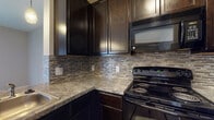 Located at 5451 Olivia Michal Pl in Westerville, Ohio, this single-floor unit offers a well-designed layout with a cohesive blend of modern finishes throughout. The kitchen serves as a functional centerpiece, showcasing rich dark espresso cabinetry, granite-style countertops, a mosaic tile backsplash, and black appliances including an electric range and over-the-range microwave. A peninsula with pendant lighting provides additional counter space and separates the kitchen from the living area, creating an open-concept feel. The transition from wood-look flooring in the kitchen to plush carpeting in the living and bedroom areas adds warmth and comfort underfoot. The living space features neutral-toned walls and access to natural light through a large sliding glass door fitted with vertical blinds, leading to a balcony. The carpeted bedrooms are generously sized, with windows dressed in horizontal blinds that allow for adjustable natural light and views of the surrounding community. The bathroom is cleanly appointed with dark-framed vanity cabinetry, white countertops, a framed mirror, and a full soaking tub and shower combination, finished with wood-look flooring for a polished look. Overall, the space presents a contemporary aesthetic with practical, well-coordinated design details throughout.
