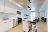 Located at 300 Mìwàte Pvt in Ottawa, this contemporary 4-bedroom, 3-bathroom unit offers a well-designed single-floor layout that balances open living with private bedroom spaces. The combined kitchen and living area serves as the heart of the home, showcasing an industrial-modern aesthetic with a distinctive exposed concrete ceiling, recessed lighting, and stylish pendant fixtures. The kitchen is outfitted with sleek white cabinetry, a marble-style backsplash, stainless steel appliances including a full refrigerator, range, and dishwasher, and a central island with bar seating — ideal for casual dining or entertaining. Light hardwood flooring flows throughout, adding warmth to the clean, minimalist palette. The living area opens toward floor-to-ceiling windows and a private balcony, offering panoramic city views and abundant natural light. Each of the four bedrooms is bright and functional, with large windows framing outdoor views. The primary bedroom includes a built-in closet and direct bathroom access. All three bathrooms are finished with large-format neutral tiles, walk-in glass showers, and modern floating vanities. A central hallway connects all spaces efficiently, rounding out this thoughtfully laid-out urban residence.