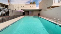This outdoor pool area presents a generously sized rectangular swimming pool filled with clear, turquoise water, offering a refreshing and inviting atmosphere. The pool features classic tiled accents along the floor and walls, along with chrome handrails for easy entry and exit. The surrounding deck is finished in a clean, light-toned concrete surface that provides ample space for lounging and movement.

Poolside amenities include bright red chairs and tables positioned near the perimeter, offering a casual seating area for guests. A covered structure adjacent to the building provides a shaded retreat, while colorful signage adds a lively visual element to the space. The enclosure is framed by white stucco walls and a black metal security fence, ensuring both privacy and safety.

Lush palm trees rise above the boundary walls, lending a warm, resort-like character to the environment. The open-air layout allows for abundant natural light throughout the day, with wide-open skies overhead enhancing the sense of openness. The overall design is functional yet inviting, combining practical poolside infrastructure with a relaxed, sun-soaked ambiance well-suited for leisure and recreation.