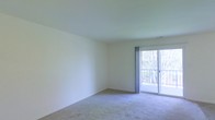 This single-floor space features a clean, minimalist living area characterized by neutral tones and a simple, functional layout. The room is finished with wall-to-wall carpeting in a soft, neutral hue that adds warmth underfoot while contributing to the overall understated aesthetic. Smooth, light-colored walls and a matching ceiling create a bright, open atmosphere throughout the space.

A prominent feature of the living area is the sliding glass door positioned along one wall, flanked by sheer curtains that filter natural light into the room. The door provides direct access to a private balcony enclosed by a metal railing, offering views of surrounding trees and greenery. This outdoor connection enhances the sense of openness and allows for ample daylight to fill the interior throughout the day.

The overall design is straightforward and versatile, presenting a blank canvas well-suited for a variety of furnishing arrangements. The combination of natural light, soft carpeting, and neutral finishes gives the space a calm, livable ambiance that is easy to personalize.