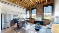 Located at 515 W 4th St in Newton, Iowa, this one-bedroom, one-bathroom loft masterfully blends industrial heritage with contemporary design. The space is defined by dramatic exposed wooden beam ceilings and original brick walls that stretch across the open-concept main living area, creating a warm, textured ambiance. Large black-framed windows flood the interior with natural light and offer views of the surrounding neighborhood. The kitchen is well-appointed with white shaker cabinetry, stainless steel appliances including a full-size refrigerator, electric range, microwave, and dishwasher, plus dark lower cabinets and a central island with seating — ideal for casual dining or entertaining. The dining and living areas flow seamlessly together beneath statement pendant lighting, with rich hardwood floors throughout. The entrance features a built-in mudroom with a wood bench, upper cabinetry, and wall hooks for organized storage. A sliding barn door adds an architectural detail that complements the industrial aesthetic. The bedroom provides a private retreat, while the bathroom offers a clean, bright finish with marble-style tile flooring, a walk-in shower, and modern black hardware accents throughout.