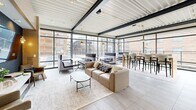 Located at 1717 Ridge Ave in Evanston, IL, this single-floor communal space offers a thoughtfully designed, open-plan layout that blends comfort and contemporary style. The space is defined by striking floor-to-ceiling glass curtain walls that flood the interior with natural light and provide sweeping views of the surrounding outdoor terrace and neighborhood beyond.

The living area is generously proportioned, featuring multiple seating arrangements including plush sofas, accent chairs in muted teal tones, and modular lounge furniture set atop layered area rugs over large-format tile flooring. A dedicated media wall with textured dark tile, wall-mounted display, and warm sconce lighting anchors one end of the room, while a work nook with café-style seating offers a more focused environment.

The kitchen and dining area is equally well-appointed, centered around a substantial white waterfall island with leather-upholstered barstools. Sage green lower cabinetry, open wood shelving, white subway tile backsplash, and globe pendant lighting lend the kitchen a warm, modern character. Large glass doors connect seamlessly to an outdoor patio.

A clean, carpeted hallway with recessed skylights and crisp white walls connects the main areas. Throughout, exposed steel beams and corrugated metal ceilings add an industrial edge, balancing the space's warm and refined furnishings.