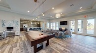 This single-story clubhouse is thoughtfully designed to offer a variety of amenity spaces within an open, cohesive layout unified by wood-look plank flooring, crown molding, and a neutral coastal color palette throughout.

The central gathering area is anchored by a full-size pool table beneath a cluster of pendant lights, with ample surrounding space for socializing. Adjacent to this is a comfortable lounge featuring plush seating, a wall-mounted television, and multiple sets of French doors that open to an outdoor patio, flooding the interior with natural light and views of the surrounding community.

The kitchen area is a standout feature, showcasing a large white quartz island with bar-height seating, a subway tile backsplash with open shelving, stainless steel appliances, and dark cabinetry—all illuminated by pendant lighting above.

A welcoming reception lobby provides additional seating and a dedicated workspace, while glass-paneled doors lead to private offices. A fitness area, visible through interior windows, includes cardio equipment, medicine balls, and direct outdoor access, completing this well-appointed community amenity space.