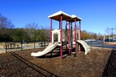 This outdoor amenity space is thoughtfully designed to serve the recreational needs of a residential community. The grounds are divided into distinct functional zones, each enclosed by sturdy black metal fencing that provides clear boundaries while maintaining an open, welcoming feel.

A dedicated dog park occupies a generous portion of the space, featuring a wood chip ground covering that provides a natural, low-maintenance surface. The area is equipped with pet amenity stations and offers ample room for dogs to roam freely. The enclosure is accessed through a secure gate, ensuring the safety of pets within.

Adjacent to the dog park is a children's playground equipped with a multi-level play structure. The unit features slides, climbing elements, and is covered by a colorful canopy overhead, all set atop a protective wood chip base. The playground is similarly enclosed by the perimeter fencing, creating a safe play environment.

The surrounding landscape is framed by a mix of deciduous and evergreen trees, offering a natural backdrop throughout the seasons. The space enjoys abundant natural light under open skies, with views of the broader residential community — including multi-story buildings with balconies and well-maintained parking areas — visible from within. The overall setting is clean, functional, and designed with community use in mind.