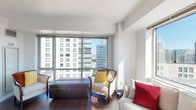 This single-floor residence offers a thoughtfully arranged layout comprising a living area, bedroom, kitchen, and full bathroom. The living room is a standout space, defined by floor-to-ceiling windows and large glass panels that frame sweeping urban skyline views, flooding the room with abundant natural light. Dark hardwood floors run throughout, providing a rich contrast to the clean white walls and ceilings.

The kitchen features a practical galley-style design with warm wood cabinetry, stainless steel appliances including a refrigerator, stove, and built-in microwave, along with a light-toned countertop that doubles as a breakfast bar. A window above the counter allows additional natural light into the space.

The bedroom is generously proportioned, accommodating a queen-sized bed with room to spare. A large picture window overlooks the cityscape, continuing the residence's emphasis on views and natural light. The room includes ample space for additional furnishings such as a dresser and media console.

The bathroom features light tile flooring, a wood-toned vanity with integrated sink, and a full shower-tub combination. The overall ambiance of the residence is modern, airy, and urban, benefiting from its elevated position with panoramic city views.