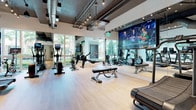 This contemporary fitness center presents a thoughtfully designed, single-level layout that balances open workout space with distinct functional zones. The main cardio area is expansive and airy, featuring light wood-tone flooring throughout and floor-to-ceiling glass windows that invite abundant natural light and offer views of surrounding greenery, creating an energizing atmosphere. The exposed ceiling with industrial-style ductwork and pendant track lighting adds a modern, loft-like character to the space.

The facility is equipped with a comprehensive selection of cardio machines, including treadmills, ellipticals, rowing machines, and stationary bikes, arranged to maximize space and flow. A large-format display screen is prominently mounted on one wall, allowing users to stay entertained or connected during workouts. Full-length mirrors line several walls, enhancing the sense of depth and allowing for form monitoring.

A separate, glass-enclosed zone houses additional strength training and functional fitness equipment, including a cable machine and free weights, providing a more focused workout environment. The overall design is clean, minimalist, and well-organized, reinforcing a professional, high-end fitness experience throughout.
