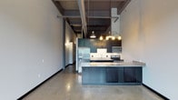 This single-floor space showcases a striking industrial-modern aesthetic that is immediately evident upon entry. The open-concept layout centers around a well-appointed kitchen, where deep teal cabinetry pairs elegantly with light granite countertops, creating a bold yet refined contrast. A generous kitchen island extends the prep and dining space, accented by stylish pendant lighting that adds warm illumination to the area. Stainless steel appliances, including a French door refrigerator with ice maker, a microwave, and a range, complete the functional kitchen suite. Polished concrete floors run throughout, lending a sleek, contemporary feel underfoot while reflecting light across the open space. The soaring ceilings draw the eye upward, where exposed ductwork and industrial-style pipes reinforce the raw, urban character of the space. Neutral gray walls provide a versatile backdrop that complements the bold design choices throughout. A corridor leads toward additional areas of the unit, hinting at further living space beyond the main open area. The combination of industrial elements with modern finishes creates an ambiance that is both edgy and inviting, making this a distinctive and thoughtfully designed space.