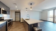 This contemporary single-floor residence is thoughtfully designed with a clean, modern aesthetic throughout. The open-concept layout centers around a standout kitchen, anchored by a generous white quartz island with a stainless steel undermount sink and a sleek industrial-style pull-down faucet. Pendant globe lights hang above the island, adding a stylish touch. Dark cabinetry lines the kitchen walls, complemented by stainless steel appliances including a French door refrigerator, gas range, and built-in microwave. Warm wood-look flooring flows seamlessly through the kitchen and into the adjacent living and dining areas, enhancing the cohesive, open feel of the space. A dedicated pantry with open shelving provides convenient storage just steps from the kitchen. The living area connects naturally to the rest of the unit, with dark-framed doorways adding a bold architectural contrast to the bright white walls. The bedroom is generously sized, bathed in natural light from a large window offering outdoor views, and features the same warm wood-look flooring. In-unit laundry is also present, adding everyday convenience. The bathroom is finished with a full tub-shower combo accented by floor-to-ceiling wood-toned tile surround, light wood-look flooring, and clean white fixtures, delivering a polished, spa-inspired feel.