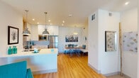 This thoughtfully designed single-floor residence offers a seamless open-concept layout that connects the kitchen, dining, and living areas into one cohesive, light-filled space. The kitchen is fully equipped with stainless steel appliances, including a refrigerator, range with a sleek overhead hood, and built-in microwave. Crisp white cabinetry with brushed hardware lines the walls, complemented by light countertops and warm hardwood flooring that flows throughout the main living areas. A central island provides additional prep space and casual seating. Adjacent to the kitchen, the dining nook features a banquette-style booth and a modern pendant light, creating an intimate yet functional setting. The living room is inviting and airy, furnished with a plush sofa, teal accent chairs, a ceiling fan, and a shag area rug, all beneath recessed lighting. The primary bedroom is a calm retreat with plush carpeting, dual windows with plantation shutters, a ceiling fan, a walk-in closet, and a wall-mounted television. The bathroom is finished in all-white with a backlit vanity mirror, undermount sink, and clean tile work, maintaining the residence's bright and modern aesthetic throughout.