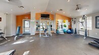 Located at 1155 W Grove Pkwy in Tempe, Arizona, this fitness center offers a well-organized, single-floor layout designed to support a comprehensive workout experience. The main gym area features vaulted ceilings with recessed lighting, creating an open and energizing atmosphere. Bold orange accent walls contrast with neutral tones throughout, adding a vibrant and motivating character to the space.

The cardio section is equipped with multiple treadmills, elliptical machines, and stationary cycling bikes, all positioned to take advantage of natural light streaming through large windows and glass double doors that open to the outdoor area. Full-length mirrors along the walls enhance the sense of space and allow for form monitoring during exercise.

The strength training area is well-appointed with a Smith machine, weight bench, cable crossover machine, medicine balls, resistance bands, stability balls, and additional free-weight equipment. A wall-mounted flat-screen television adds convenience for entertainment during workouts.

The facility also includes two utility rooms supporting the overall operation of the space. Durable rubber and concrete flooring throughout the gym provides both safety and easy maintenance. The combination of natural light, varied equipment, and thoughtful layout makes this fitness center a functional and inviting amenity.