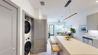 This thoughtfully designed 1-bedroom, 1-bathroom residence offers a cohesive, modern aesthetic across its single level. The open-concept kitchen and living area serves as the heart of the home, where a spacious white quartz island with bar seating anchors the space beneath a sleek linear pendant light. The kitchen features sage-toned shaker cabinetry, a white subway tile backsplash, stainless steel appliances, and light wood-look flooring that flows throughout the entire unit. A stacked in-unit washer and dryer is conveniently integrated near the entryway. The living room is bright and inviting, benefiting from large windows and a sliding glass door that welcome natural light and outdoor views, complemented by a ceiling fan overhead. The bedroom is a calm, well-lit retreat with a ceiling fan, a large window with sheer curtains, and light wood flooring. The bathroom is thoughtfully laid out with a long vanity featuring gray shaker cabinetry, white countertops, and direct access to the dedicated closet. Clean white walls, consistent flooring, and a neutral color palette lend the entire space a polished, cohesive feel.