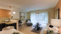 This well-appointed single-floor residence offers a thoughtfully designed open-concept layout that connects the living and kitchen areas in a cohesive, functional flow. The living area is a welcoming focal point, centered around a sleek dark coffee table set atop a textured area rug, complemented by a contemporary arc floor lamp that adds both style and ambient lighting. Large sliding glass doors dressed with vertical blinds flood the space with abundant natural light and provide views of lush greenery beyond, creating a seamless connection between the interior and the outdoors. A comfortable chaise lounge is positioned to take advantage of the natural light, adding a relaxed, refined touch to the room. The kitchen area features a raised breakfast bar with modern bar seating and a stylish pendant light overhead, offering a casual dining option distinct from the main living space. Light hardwood-style flooring runs throughout, contributing to the airy and cohesive aesthetic. Warm, neutral wall tones are accented by a vibrant piece of framed abstract artwork, lending a splash of color and personality to the space. The overall ambiance is modern, clean, and inviting, blending comfort with contemporary design.