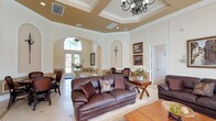 This sophisticated single-floor space exudes classic elegance through its thoughtfully designed interiors and refined architectural details. The expansive main lounge serves as the centerpiece, showcasing soaring tray ceilings adorned with decorative crown molding and warm contrasting paint, illuminated by ornate wrought-iron chandeliers. The walls feature raised panel wainscoting with arched niches and framed artwork, contributing to a polished, formal ambiance throughout.

Rich, dark leather seating arrangements are complemented by warm wood accent tables, creating inviting conversation areas across the generous open floor plan. Large French doors dressed in flowing gold and champagne draperies line an entire wall, flooding the space with natural light while offering direct views of a sparkling outdoor pool. A dedicated dining area with dark wood chairs and a round table provides an ideal setting for gatherings.

Light-toned porcelain tile flooring with decorative inlay patterns extends seamlessly throughout, enhancing the sense of spaciousness. The functional kitchen features rich cherry wood cabinetry, stone countertops, stainless steel appliances, and a window that brings in additional natural light. Together, these elements create a space that is both grand in scale and warm in character, well-suited for entertaining and community gatherings alike.