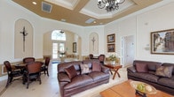 This sophisticated single-floor space exudes classic elegance through its thoughtfully designed interiors and refined architectural details. The expansive main lounge serves as the centerpiece, showcasing soaring tray ceilings adorned with decorative crown molding and warm contrasting paint, illuminated by ornate wrought-iron chandeliers. The walls feature raised panel wainscoting with arched niches and framed artwork, contributing to a polished, formal ambiance throughout.

Rich, dark leather seating arrangements are complemented by warm wood accent tables, creating inviting conversation areas across the generous open floor plan. Large French doors dressed in flowing gold and champagne draperies line an entire wall, flooding the space with natural light while offering direct views of a sparkling outdoor pool. A dedicated dining area with dark wood chairs and a round table provides an ideal setting for gatherings.

Light-toned porcelain tile flooring with decorative inlay patterns extends seamlessly throughout, enhancing the sense of spaciousness. The functional kitchen features rich cherry wood cabinetry, stone countertops, stainless steel appliances, and a window that brings in additional natural light. Together, these elements create a space that is both grand in scale and warm in character, well-suited for entertaining and community gatherings alike.