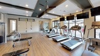 This fitness center offers a comprehensive and thoughtfully designed workout environment spread across a single open floor. The space is finished with warm hardwood-style flooring and features a coffered ceiling with decorative dark wood beams, recessed lighting, and ceiling fans that contribute to a comfortable exercise atmosphere.

The cardio zone is generously outfitted with multiple treadmills and elliptical machines, each equipped with individual mounted flat-screen televisions for entertainment during workouts. Large floor-to-ceiling windows and glass doors flood the area with natural light and provide views of an outdoor patio, creating an open and energizing environment.

The strength training area includes a full rack of dumbbells in varying weights, a multi-functional cable machine, and additional resistance equipment to support a wide range of exercises. An adjustable bench and a dedicated TRX suspension training system round out the functional fitness options. Full-length mirrors are strategically placed to expand the sense of space and allow users to monitor their form.

A decorative accent wall featuring a geometric, cross-shaped panel design in muted sage green and warm neutral tones adds a distinctive visual element to the space. The overall ambiance is modern and welcoming, balancing practical fitness functionality with clean, stylish interior design.