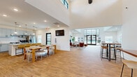 This well-appointed single-story community space offers a generous and thoughtfully designed layout across its open floor plan. The centerpiece is a dramatic open-concept area combining the dining room, living room, and kitchen into one expansive, flowing space. Soaring double-height ceilings amplify the sense of openness, while large black-framed grid windows and glass-paneled entry doors flood the interior with abundant natural light and provide views of the surrounding outdoor landscape.

The living area features warm wood-look tile flooring throughout, a cozy seating arrangement anchored by a fireplace with a wood beam mantel, and a wall-mounted television. A shuffleboard table adds a recreational element to the lounge zone. The dining area accommodates a long communal table ideal for gatherings, complemented by bar-height bistro tables nearby.

The kitchen is outfitted with light gray shaker cabinetry, granite countertops, stainless steel appliances including a range, dishwasher, and French door refrigerator, and a central island with pendant globe lighting above. A dedicated office nook provides two individual workstations, each featuring wall-mounted shelving, compact desks, and comfortable seating. A hallway connects the various zones, completing this versatile and welcoming community space.