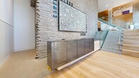Located at 1515 Surf Ave in Brooklyn's Coney Island neighborhood, this two-floor space offers a combined footprint across both levels, making it a substantial and versatile property. The ground floor serves as an impressive lobby and gathering area, immediately drawing attention with its textured stacked-stone accent wall adorned with large-scale Coney Island-themed artwork. Light hardwood flooring runs throughout, complementing the warm wood-paneled ceiling elements and exposed concrete columns that define the architectural character of the space.

Floor-to-ceiling glass walls and double-door entries flood the ground level with natural light, framing street views and creating a seamless connection to the outdoors. A prominent feature of this floor is a wide, tiered staircase dressed with colorful cushions, functioning both as a circulation element and a casual gathering amphitheater. Glass-railed staircases with brushed metal handrails provide elegant transitions between floors.

The upper level continues the refined aesthetic with open sightlines, glass balustrades overlooking the lobby below, and access to additional functional areas. A secured hallway with smart-access technology and dedicated mailbox stations adds a layer of modern convenience. The overall ambiance is contemporary and vibrant, blending industrial materials with warm finishes to create an inviting, design-forward environment.
