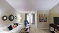This single-floor residence offers a thoughtfully designed living space that blends comfort with contemporary style. The living room serves as the heart of the home, showcasing plush wall-to-wall carpeting that adds warmth underfoot. A vaulted ceiling enhances the sense of openness, while a ceiling fan provides both function and a finishing architectural touch. Natural light filters generously into the room through a set of elegant French doors dressed with floor-length drapes, offering a seamless connection to the outdoors. The space is tastefully appointed with a clean white sofa accented by bold orange and teal throw pillows, a tufted navy accent chair, and a rustic wood coffee table that grounds the seating arrangement. A vibrant geometric canvas on one wall introduces a lively pop of color, complementing the neutral beige tones of the walls and flooring throughout. A dedicated corridor leads to additional areas of the home, suggesting a well-organized and functional floor plan. The overall ambiance is light, airy, and inviting — ideal for both relaxed everyday living and comfortable entertaining.